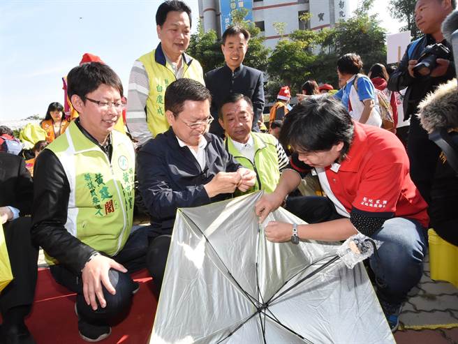 彰化縣長魏明谷(左二)做在小板凳上與民眾一童體驗手工縫傘，看來頗耗費眼力。(謝瓊雲攝)