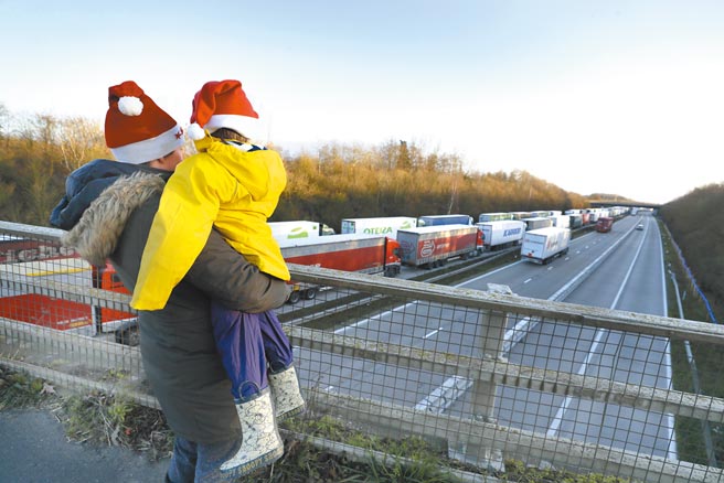 一位母亲25日抱着小孩在英国肯特郡阿什福德M20高速公路陆桥上，观看卡车大排长龙驶向多佛港检查站，准备乘渡轮穿越英吉利海峡前往法国加来港。英国出现变种新冠病毒，导致欧洲多国关闭与英国的交通联繫，从而「预演」英国无协议脱欧可能出现的乱象。（美联社）