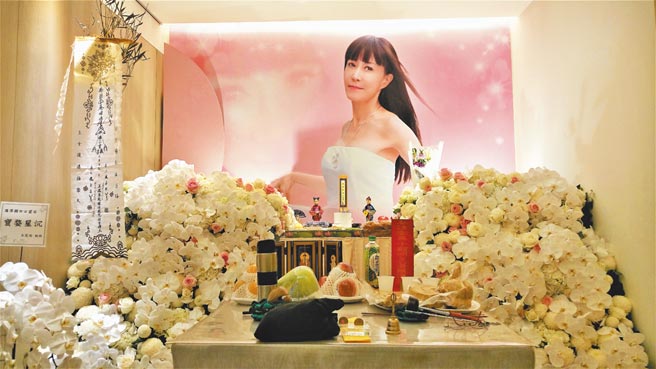 高桌两旁，放满白菊花与白蝴蝶兰，中间点缀浅淡粉红玫瑰，配合整墙的粉红背景，素雅温馨之感，瀰漫整个灵堂。（罗青提供）