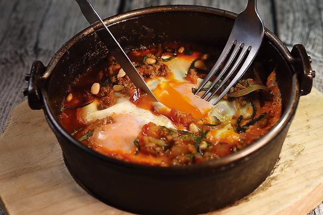 〈夏卡蘇卡〉是用小鐵鍋將大量番茄、洋蔥、辣椒、大蒜炒香，再加入摩洛哥香料與番茄混合，最後打入兩顆水波蛋，撒上烤至香酥的松子及杏仁片。圖／姚舜