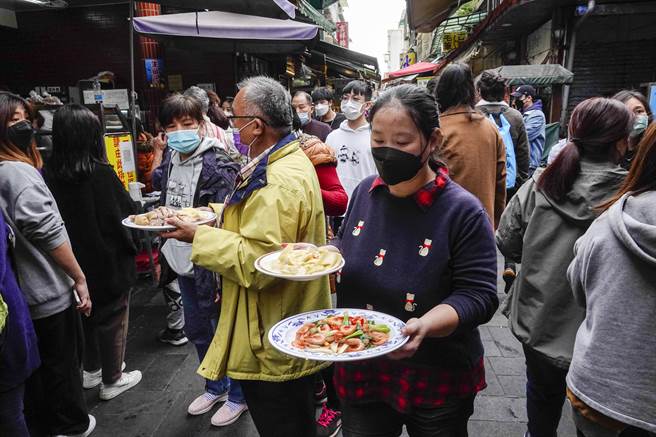 金山老街挤满人潮，民眾涌入知名美食店家品尝佳肴，由于店家厨房与餐席分离，饕客端着菜肴穿梭人群中前往老街多处店面享用。（黄子明摄）