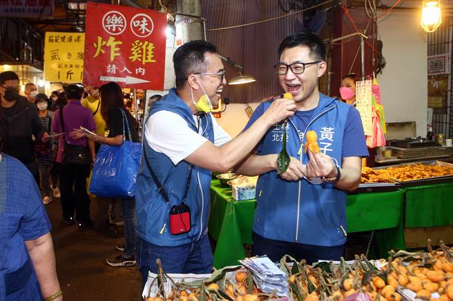國民黨立法委員江啟臣（右）10日陪同黃子哲（右2）、詹為元在士北、松信聯合拜票，江啟臣在士林華榮市場掃街拜票時，拿起琵琶教現場民眾怎麼撥琵琶，然後與黃子哲兩人相互餵食琵琶。（鄧博仁攝）