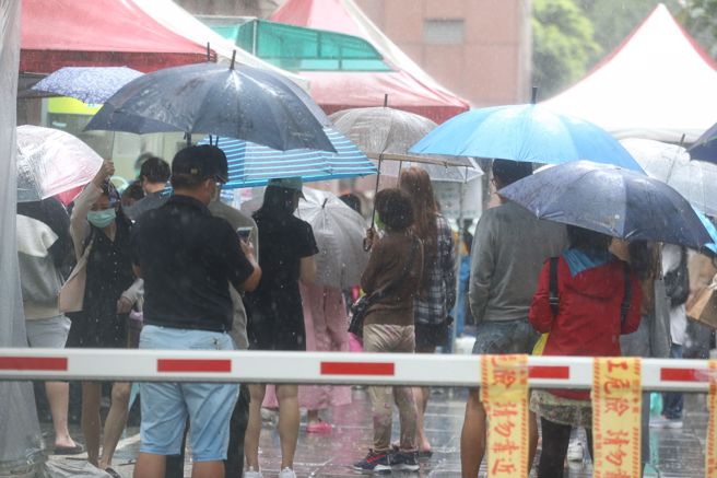 疫情严峻导致医疗量能吃紧，许多民眾在大雨滂沱中撑着雨伞等待看诊。（杜宜谙摄）