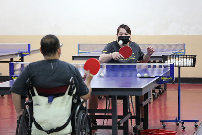 為了與身障者維持同樣高度，蔡貴蘭教練坐在椅子上陪伴肢體障礙者桌球推廣營的學員練球。(攝影/Carter)

