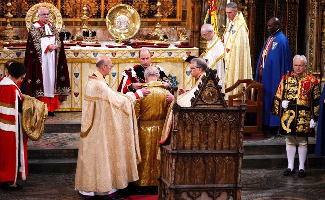 英王查尔斯6日在西敏寺内跪受恩膏，金袍加身。长子威尔斯亲王威廉替查尔斯披上长围巾。（路透）
