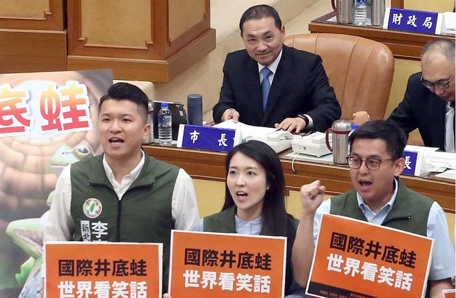新北市长侯友宜（后）9日出席新北市议会第4届第1次定期会议备询，新北市民进党市议会党团议员们手拿标语讽刺他是井底之蛙，缺乏世界观。（赵双杰摄）