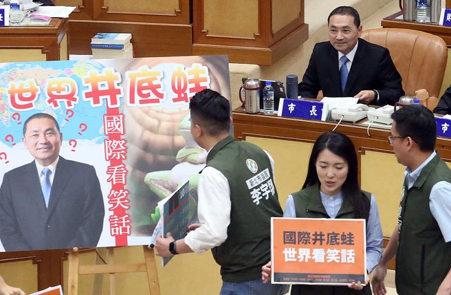 新北市长侯友宜（后）9日出席新北市议会第4届第1次定期会议备询，新北市民进党市议会党团议员们手拿标语讽刺他是井底之蛙，缺乏世界观。（赵双杰摄）