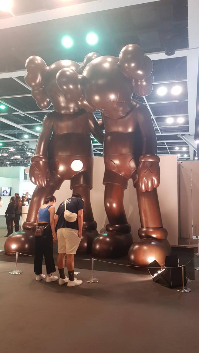 图二：佳士得香港春拍陈列KAWS高达5.5公尺的《ALONG THE WAY》，两位参观者低头查看解说，与作品姿态相同，别具趣味。（陶福媛摄）