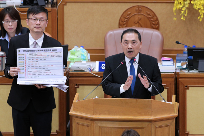 新北市幼儿园喂药案引起外界瞩目，新北市议会6日变更13日的大会议程，加开1小时请教育局及卫生局专案报告，新北市长侯友宜（右）13日赴市议会报告，教育局长张明文（左）在一旁举牌，强调市府没有消极处理。（黄世麒摄）