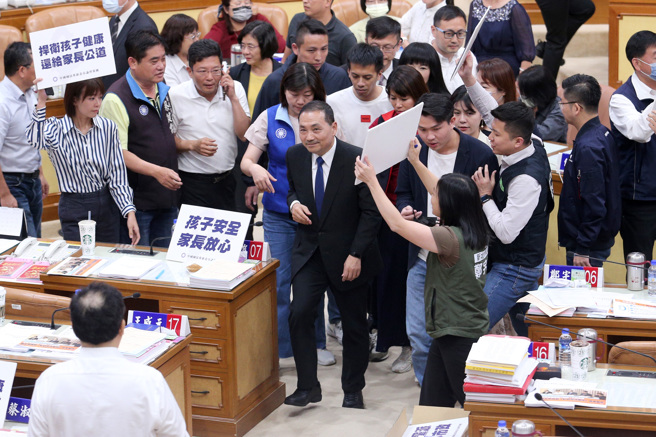新北市板桥私立幼儿园喂药案引起外界瞩目，新北市议会6日变更13日的大会议程，加开1小时请教育局及卫生局专案报告，新北市长侯友宜（中）13日赴市议会报告结束后，民进党新北市议员们不满侯市府避重就轻，上前举牌抗议，侯友宜在国民党新北市议员的保护下快步离开议会。（黄世麒摄）