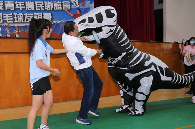新北市长侯友宜（中）16日出席青年农民联谊会羽毛球友谊赛，并担任开球嘉宾，主办单位安排一只恐龙装扮的吉祥物与侯友宜互动，结果恐龙的嘴巴不慎直接撞上侯的脸，侯友宜见状不以为意，哈哈大笑。（黄世麒摄）