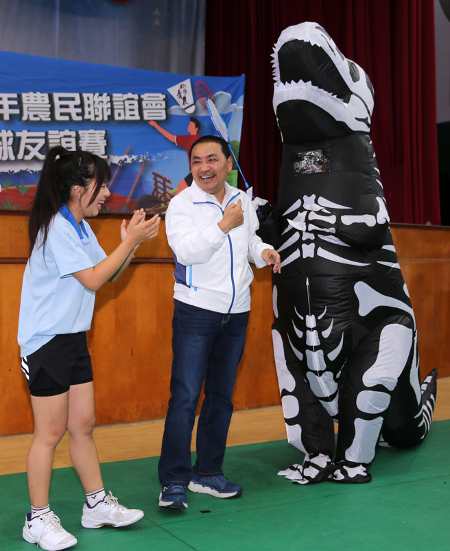 新北市长侯友宜（中）16日出席青年农民联谊会羽毛球友谊赛，并担任开球嘉宾，主办单位安排一只恐龙装扮的吉祥物与侯友宜互动，结果恐龙的嘴巴不慎直接撞上侯的脸，侯友宜见状不以为意，哈哈大笑。（黄世麒摄）