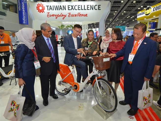 马来西亚台湾形象展今吉隆坡登场，贸协董事长黄志芳(中)于台湾精品馆体验向该国贵宾谢介绍共享经济微笑单车。(贸协提供)
