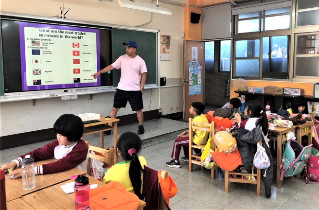 国际见习生Jeff(讲台上)带领中湖国小的学生于课堂中认识各国货币。(照片/新北市政府教育局提供)