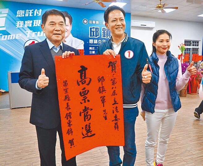 苗栗縣二選區候選人邱鎮軍（左二）得票數一路領先對手曾玟學，他13日晚間6點多在競選總部由妻子詹美娟（右一）陪同自行宣布當選，縣長鍾東錦（左一）到場致贈紅榜恭賀。（李京昇攝）