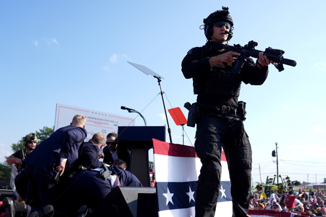 美国前总统川普13日造势遭到枪击受伤，现场画面显示，一名身穿沙漠迷彩装的白人枪手躲在附近屋顶开枪，但在川普右方屋顶的特勤局狙击手疑似早已发现枪手身影，枪手一开枪后，狙击手立刻还击将他击毙。图为川普受伤趴下后，特勤人员围在他身边保护。（美联社）