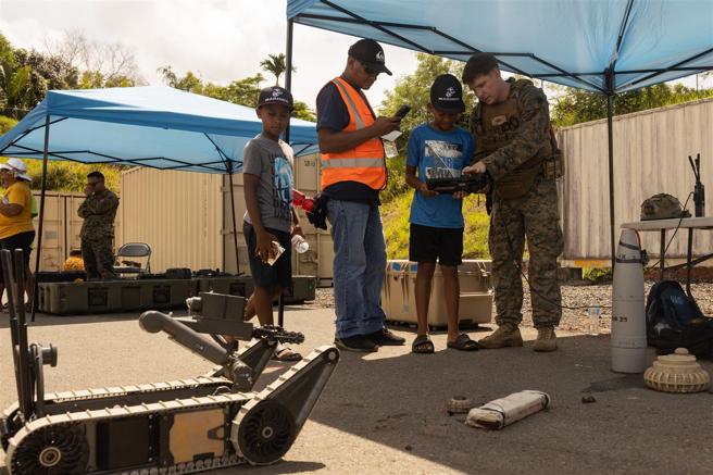 孩子们在陆战队员指导下，操作遥控无人载具，模拟拆除地面障碍物。（取自DVIDS网站）